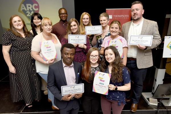 A group of happy people stand and kneel together, holding various placards and certificates